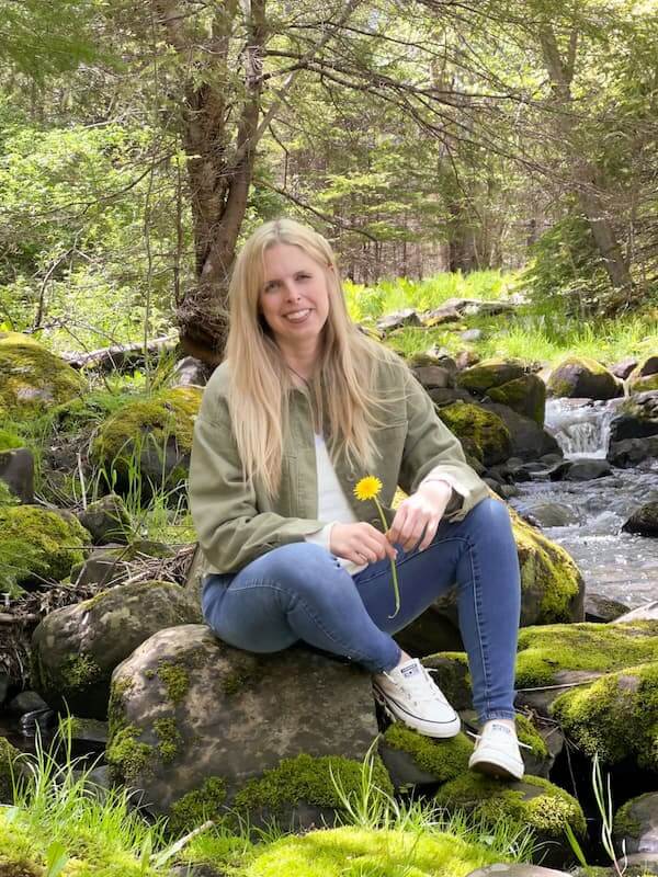 About Jill MacKay - Energy Recovery Specialist for Women, sitting on rock in the woods holding a yellow flower