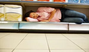 Exhausted woman asleep on a supermarket shelf, symbolizing burnout fatigue that doesn’t lift with rest.